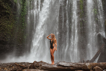 Young slim brunette woman in black swimsuit enjoying in lagoon of huge tropical waterfall Tiu Kelep on Lombok Island.  Happy vacations in Indonesia. © Yevhenii