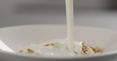 pour yogurt over granola with almond flakes and pumpkin seeds into white bowl closeup