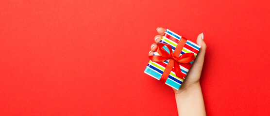 Fototapeta na wymiar Female's hands holding striped gift box with colored ribbon on red background. Christmas concept or other holiday handmade present box, concept top view with copy space