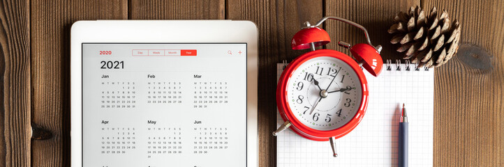 a tablet with an open calendar for 2021 year, a red alarm clock, a pine cone, and a spring notebook with a pen on a wooden boards table background. banner