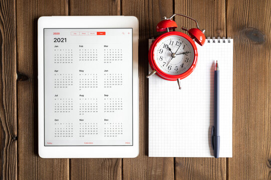 a tablet with an open calendar for 2021 year, a red alarm clock, and a spring notebook with a pen on a wooden boards table background