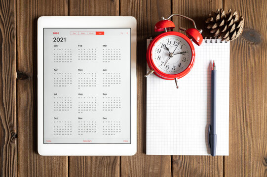 a tablet with an open calendar for 2021 year, a red alarm clock, a pine cone, and a spring notebook with a pen on a wooden boards table background
