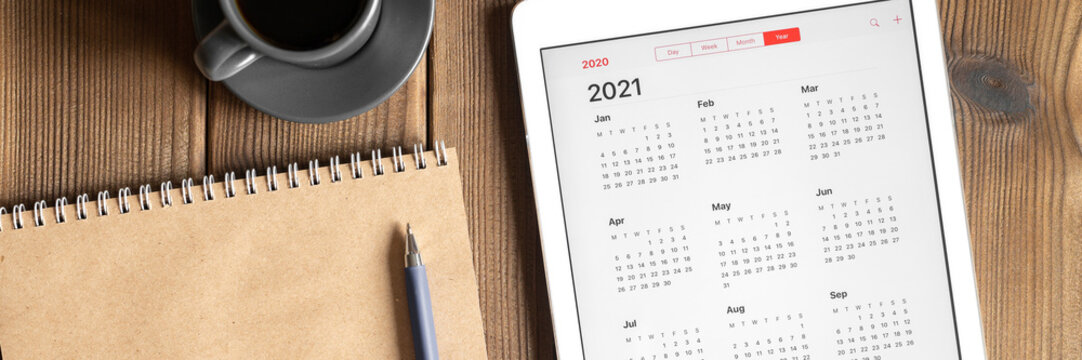 a tablet with an open calendar for 2021 year, a cup of coffee and a craft paper notebook on a wooden boards table background. banner