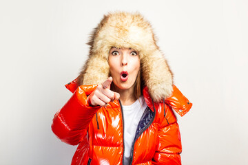 surprised young girl in a red jacket and a fur hat, looks at the camera and points a finger in front of him on a light background. Concept gestures, advertising, fashion