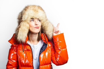young girl in a red jacket and a fur hat, looks up and points a finger up on a light background....
