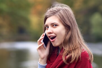 Surprised shocked happy young woman talking on cell mobile smart phone outdoors in autumn park with open mouth and excited look