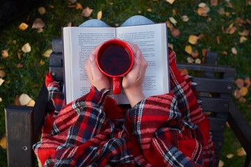 Beautiful young woman drinks tea from a red mug and reads a book outdoors in an autumn park, top view.