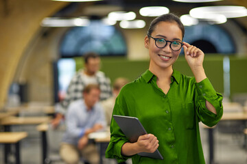 Portrait of young happy asian woman adjusting her eyeglasses, holding digital tablet and smiling at camera while standing in the office with colleagues on the background - obrazy, fototapety, plakaty
