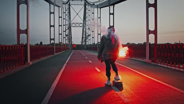Young hipster female in informal outfit is skateboarding on bridge holding burning red signal flare and waving it