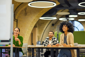 Group of young happy multiracial people sitting at desks in the modern office and listening to coach or speaker during corporate team building training or business seminar