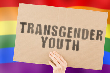 The phrase " Transgender youth " on a banner in men's hand with blurred LGBT flag on the background. Homosexual. Homosexuality. Couple. Equality. Sexual orientation. Love. Justice