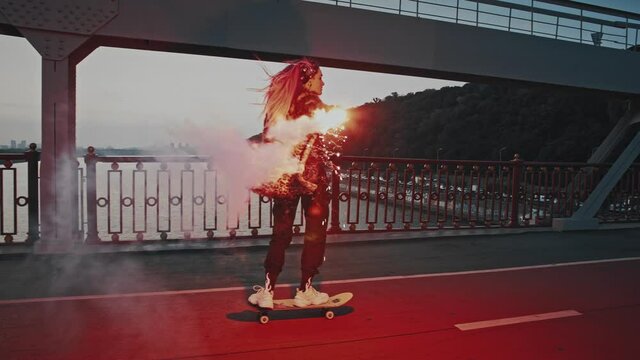 Young hipster female in informal outfit is riding skateboard on bridge holding burning red signal flare and waving it