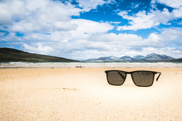 Sunglasses on sandy beach in summer - mountains, ocean and blue sky