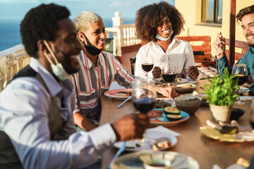 Happy multiracial friends with protective face masks eating and drinking wine together - Social distance concept - Focus on black girl's face