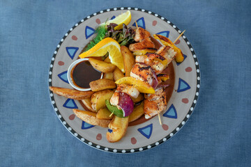Salmon and prawns with bell peppers and onions roasted on skewers, potato wedges and bbq sauce on a colorful rustic plate on a blue tablecloth, copy space, high angle view from above, selected focus
