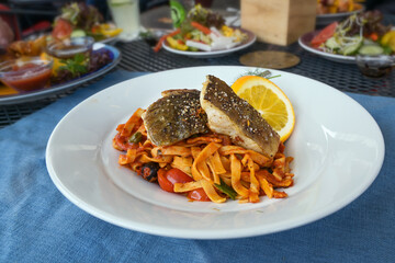 Roasted codfish on cajun pasta with red pesto, tomato and olives, orange, lemon and dill garnish, white plate and blue napkin on a table at a dinner with friends