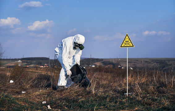 Research scientist in protective suit and gas mask picking up garbage in grassy field with warning symbol of poisonous substances and danger. Concept of ecology, environmental pollution and hazard.