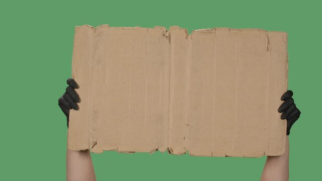 Protest banner copy space. A woman holds a blank cardboard in her hand. Advertising platform. Hand in a black medical glove isolated on a green background, chromakey. Close up. Slow motion.