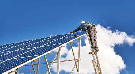 Horizontal snapshot of a male worker wearing a uniform, standing on a ladder on the top of a solar plant, mounting solar modules on a sunny day. Clear blue sky and one white cloud on background © anatoliy_gleb