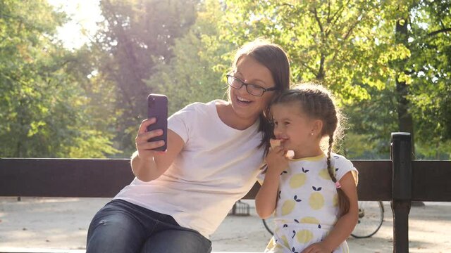 Mother takes selfie picture vie smartphone with her little child girl daughter eating ice cream on park bench in good sunny day