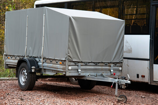 A Car Trailer Covered With A Gray Tarpaulin In An Unpaved Parking Lot.