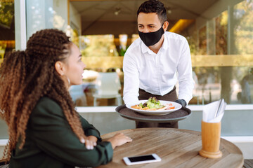 Young waiter in  protective face mask and gloves while bringing food to a customer in cafe during coronavirus epidemic. Covid- 2019.