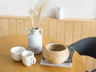cups and vase on the table. Scandinavian interior. wooden round table and wood walls. wicker basket and magazines. In the vase of dried flowers