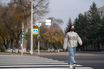A girl in a black face mask crosses the road at a pedestrian crossing at a green traffic light. Traffic Laws concept