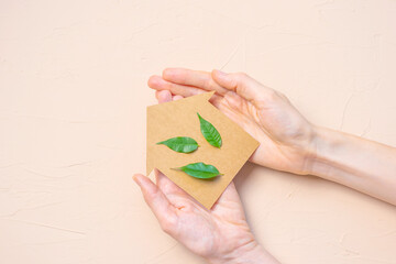 House made of craft paper and green leaves of a plant in female hands on a light background. Eco-friendly home concept, healthy lifestyle, zero waste.