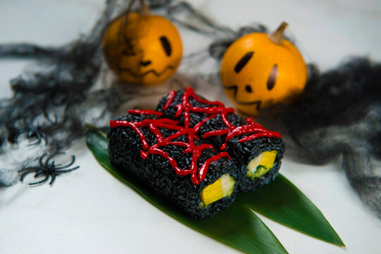 Japanese Sushi Roll With Halloween Decoration On Background. Spiders, Bat, Candle And Pumpkins With Scary Painted Faces. Sushi Roll Wrapped In Black Rice, Decorated With Red Spider Web On Top
