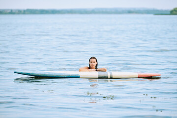 Young attractive sportswoman with dark wet hair standing in water by surfboard
