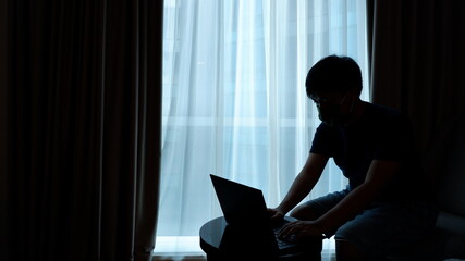 A young asian man with a mask in a silhoutte is working with his laptop in his home (work from home)