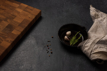 Empty black cast iron frying pan on a dark graphite background. Light linen towel. Wooden end cutting board. Fresh rosemary.