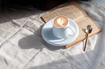 Relaxing holiday morning with hot heart-shaped latte art coffee in a white coffee mug put on the book with the warm morning sunshine from the window to feel relaxed good for relaxing