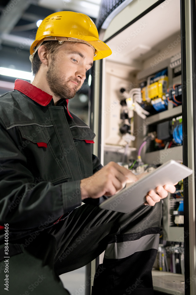 Poster young bearded engineer in workwear and hardhat checking settings of machine