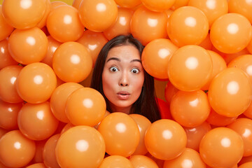 Fototapeta na wymiar Photo of shocked brunette young Asian woman sticks head through orange inflated helium balloons has stunned expression celebrates holiday poses against decorated background. Festive event concept
