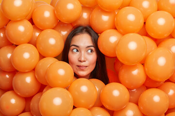 Fototapeta na wymiar Horizontal shot of dreamy thoughtful woman celebrates special occasion has long dark straight hair surrounded by many small inflated balloons thinks about party celebration enjoys favorite holiday
