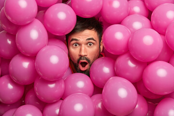 Fototapeta na wymiar Close up shot of shocked bearded European man looks with widely opened mouth at camera being greatly surprised poses over inflated pink balloons impressed to get unexpected gift on birthday.