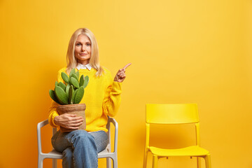 Serious middle aged woman poses with cactus at chiar looks confidently and points away on empty...