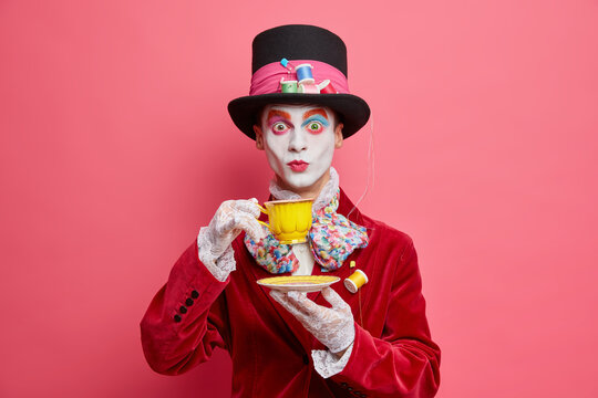 Surprised gentleman has image of character from wonderland wears aristocratic costume lace gloves hat and drinks tea has colorful skull makeup isolated on pink background. Carnival party concept