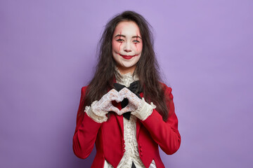 Beautiful smiling woman with fearful makeup pale ghost face and bloody scars makes heart gesture...