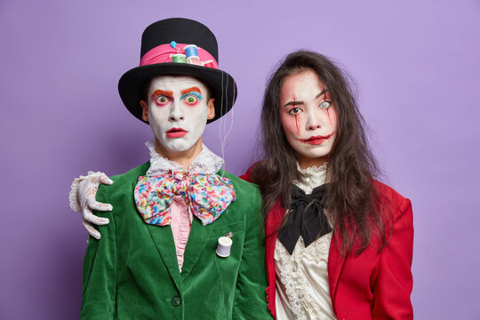 Two friends dressed in halloween carnival costumes embrace and have friendly relationships wear spooky makeup celebrate holidays isolated on purple background. Scared hatter poses with female zombie