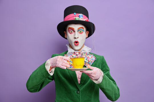 Mad Tea Party. Crazy Shocked Man Hatter Wears Large Top Hat Oversized Bowtie Lace Gloves And Green Jacket Drinks Hot Beverage Has Image Of Fictional Character Celebrates Halloween Stands Indoor