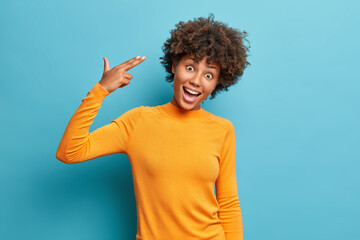 Joyful African American female foolishes around shoots in temple with fingers makes suicide gesture dressed in casual jumper poses against blue background. Funny female makes finger gun pistol