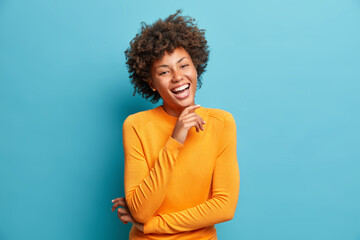 Portrait of glad young Afro American woman laughs happily keeps hand on chin expresses positive emotions smiles broadly has carefree expression wears orange jumper isolated over blue background - obrazy, fototapety, plakaty