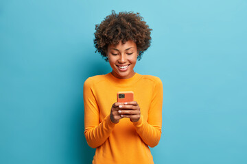 Fototapeta Online lifestyle concept. Cheerful good looking woman with Afro hair sends text messages via mobile phone dressed casually searches gifts for holiday in internet uses smartphone app browses webpage obraz