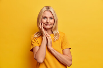 Studio shot of calm beautiful woman with healthy skin touches face gently wears minimal makeup has tender smile takes care of her complexion wears yellow t shirt in one tone with background. - obrazy, fototapety, plakaty