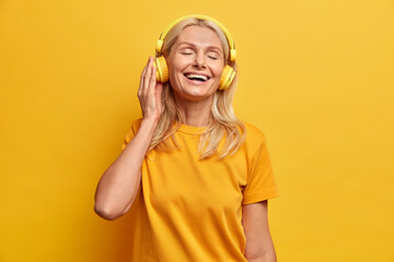 Mature blonde woman listens favorite music in headphones chooses best song from playlist enjoys favorite hobby stands with closed eyes dressed in casual t shirt poses against yellow background