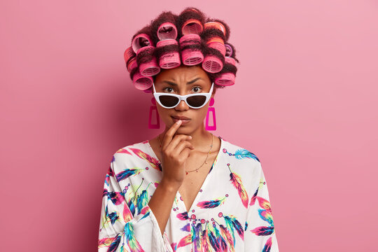 Serious strict dark skinned woman wears sunglasses and dressing gown makes curly hairstyle with hair rollers thinks about her image on party poses indoor against pink background. Beauty concept