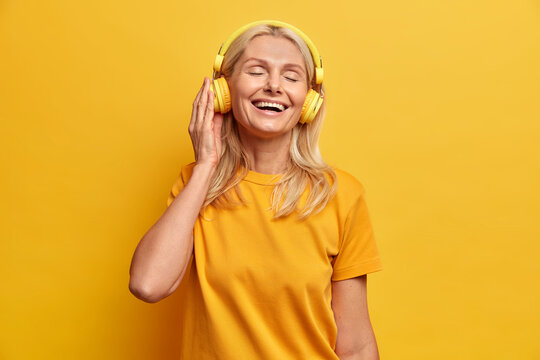 Mature Blonde Woman Listens Favorite Music In Headphones Chooses Best Song From Playlist Enjoys Favorite Hobby Stands With Closed Eyes Dressed In Casual T Shirt Poses Against Yellow Background